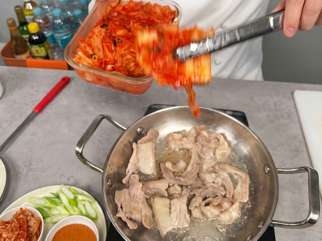Une portion de kimchi rouge vif transférée dun bocal vers la poêle contenant le porc doré.