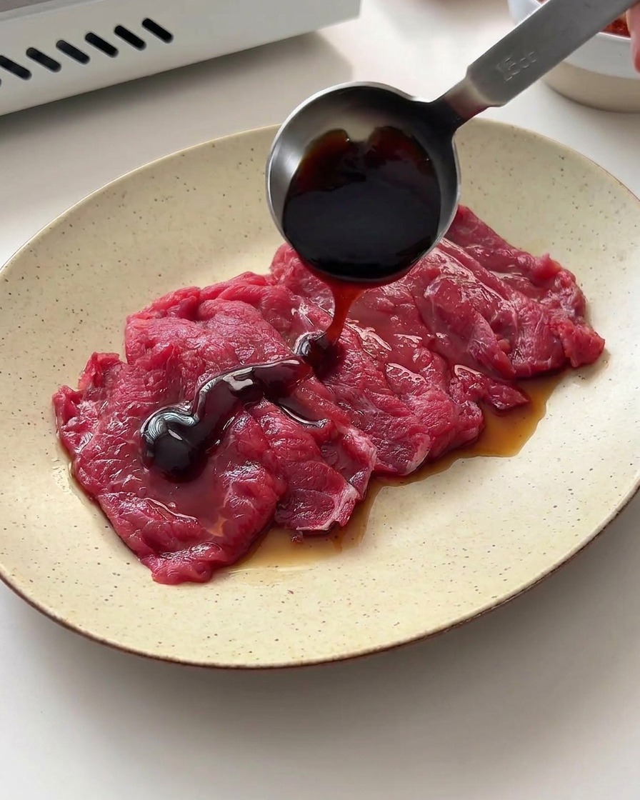 Versement de sauce soja et de sauce dhuître sur des tranches de bœuf cru à laide dune cuillère doseuse.