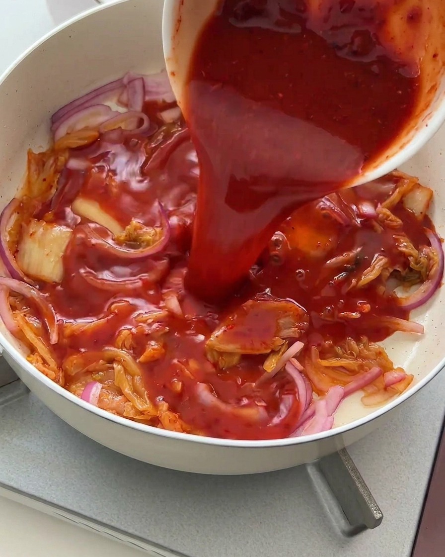Sauce rouge vibrante versée depuis un bol blanc sur le mélange de kimchi et doignons.
