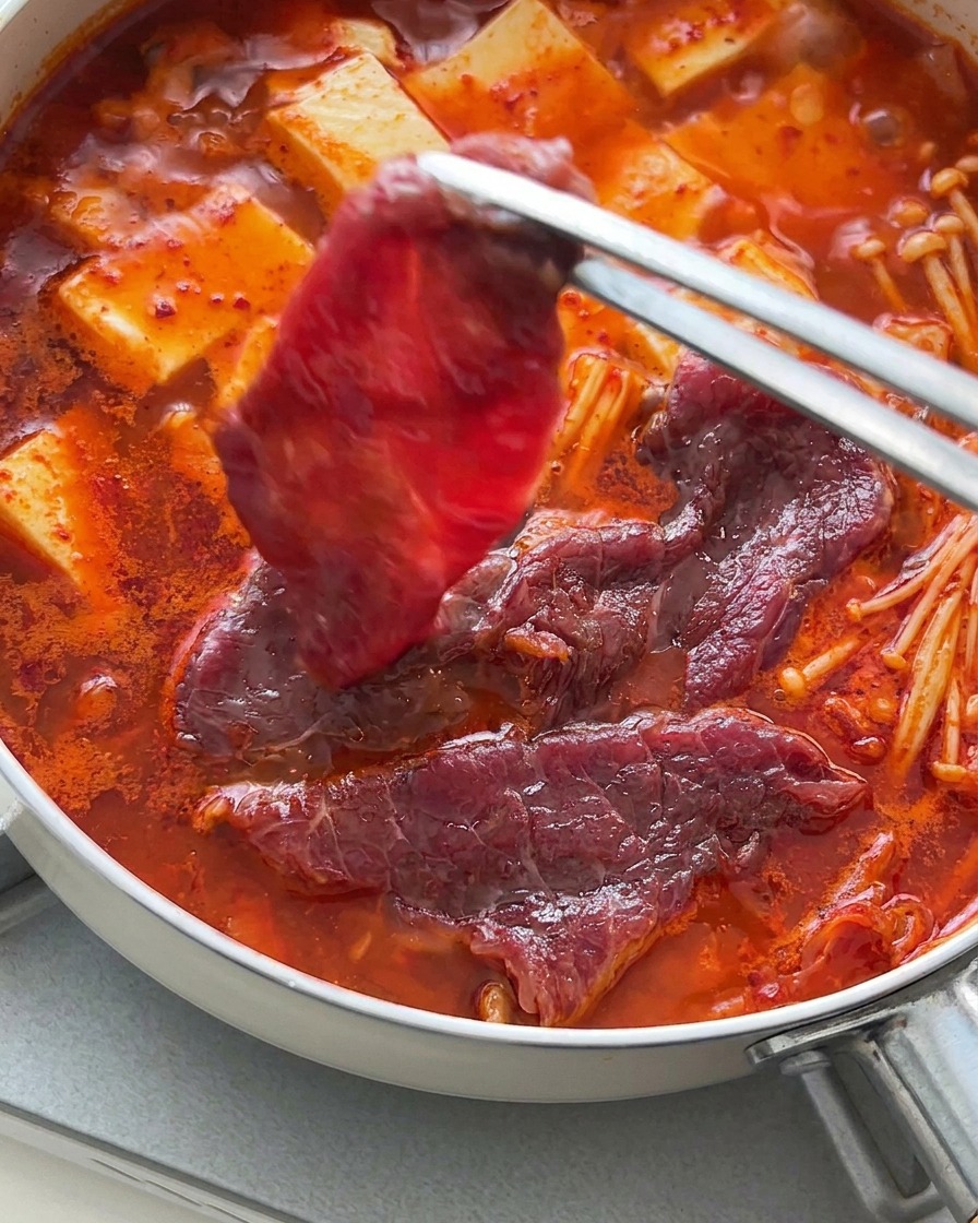 Pince disposant des tranches de bœuf cru sur le dessus du ragoût de tofu et kimchi fumant.