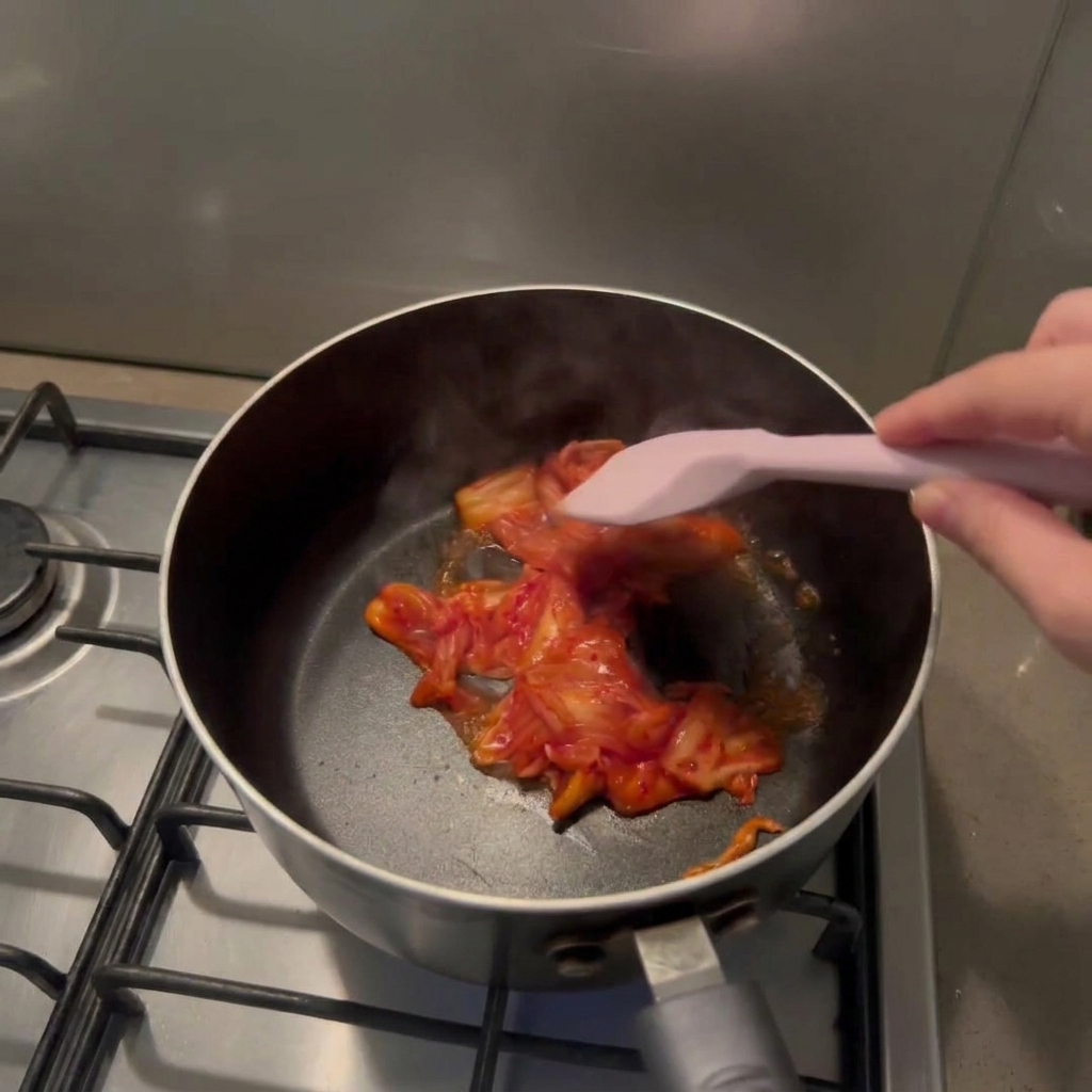 Une main utilisant une spatule en silicone rose pour faire sauter du kimchi rouge haché dans une marmite noire.