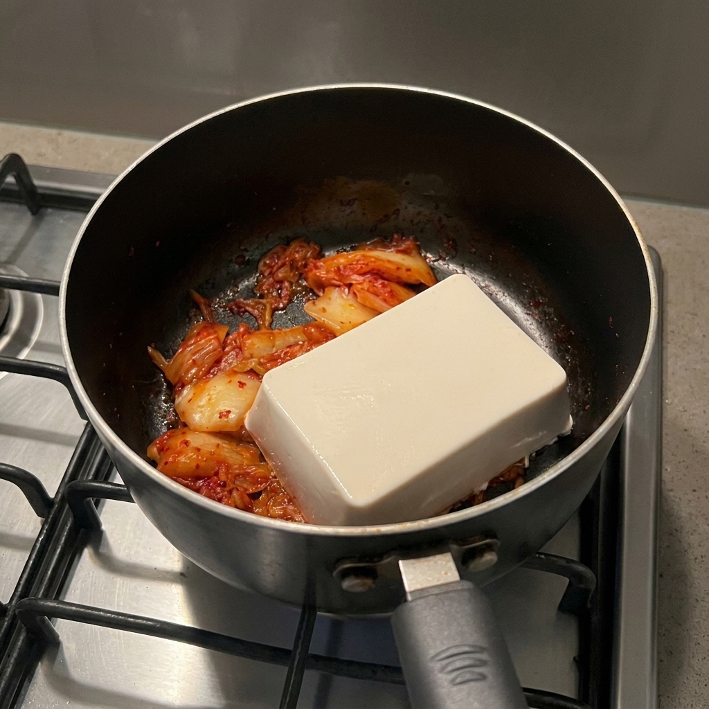 Un bloc rectangulaire de tofu soyeux blanc posé sur du kimchi rouge cuit dans une marmite noire.