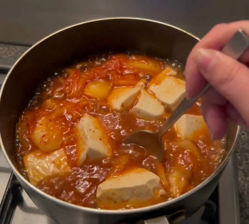 Une cuillère mélangeant de la pâte de piment dans une soupe bouillonnante contenant du tofu et du kimchi.