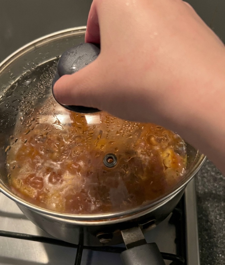 Une main tenant le bouton noir dun couvercle en verre et le posant sur une marmite de ragoût fumant.