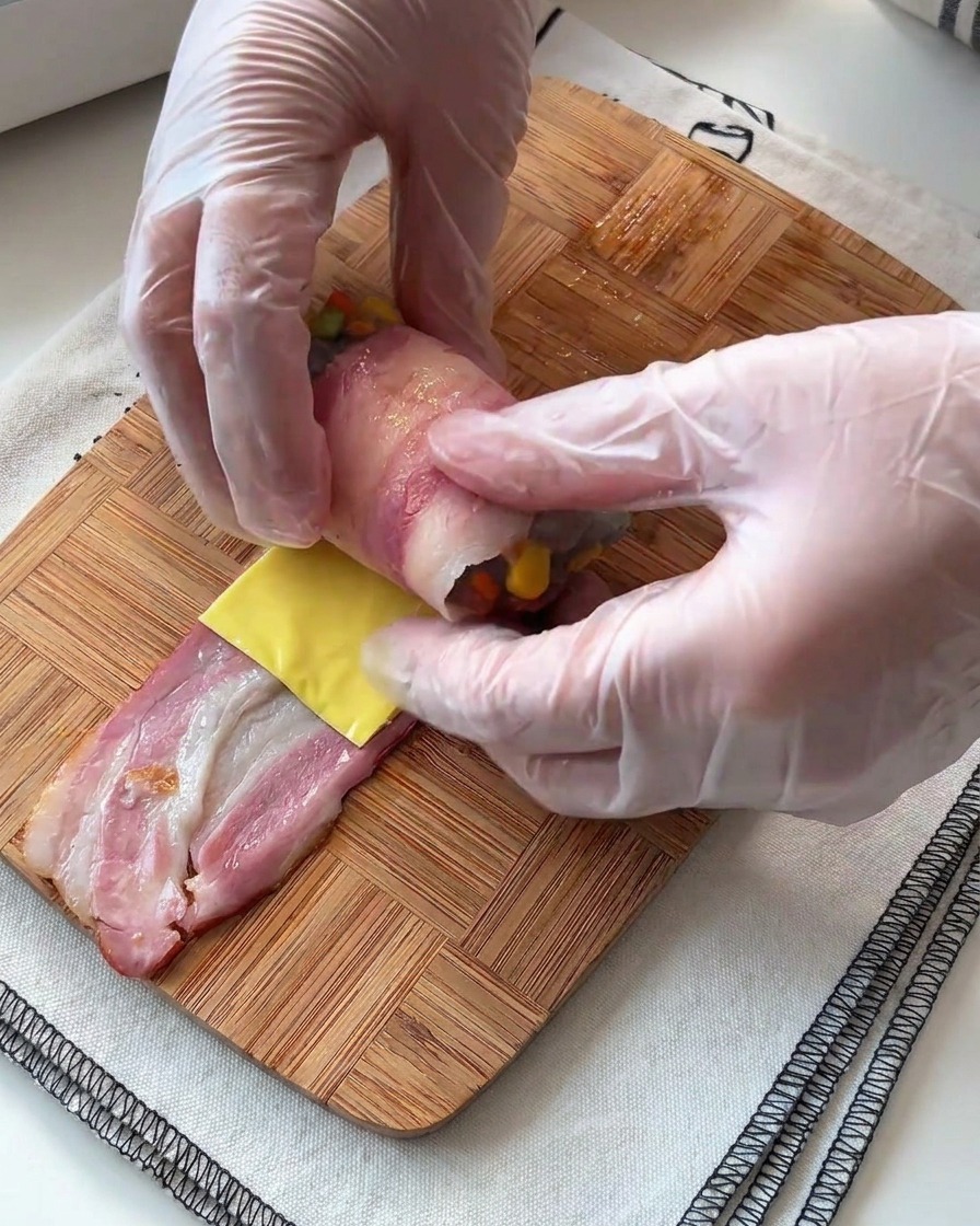 Des mains enroulant délicatement une tranche de lard autour dune farce aux crevettes et au fromage sur une surface en bois.