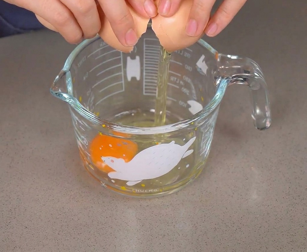 Mains cassant un œuf brun dans un verre contenant déjà un premier œuf cru.
