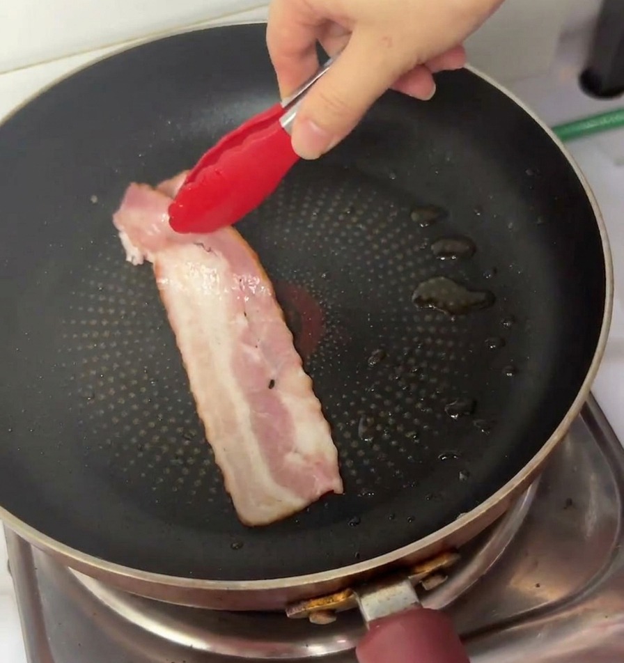 Une main utilisant des pinces pour placer une tranche de bacon cru dans une poêle antiadhésive.
