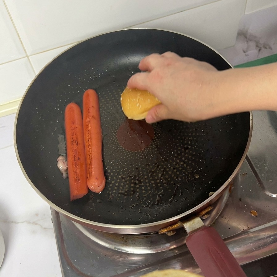 Une personne plaçant le chapeau dun pain à burger face contre la poêle à côté des saucisses grillées.