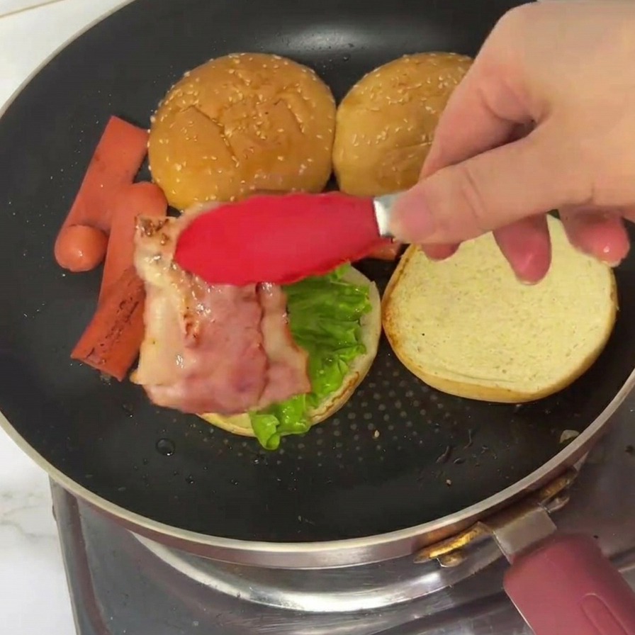 Des pinces déposant une tranche de bacon grillé sur le pain garni de laitue.