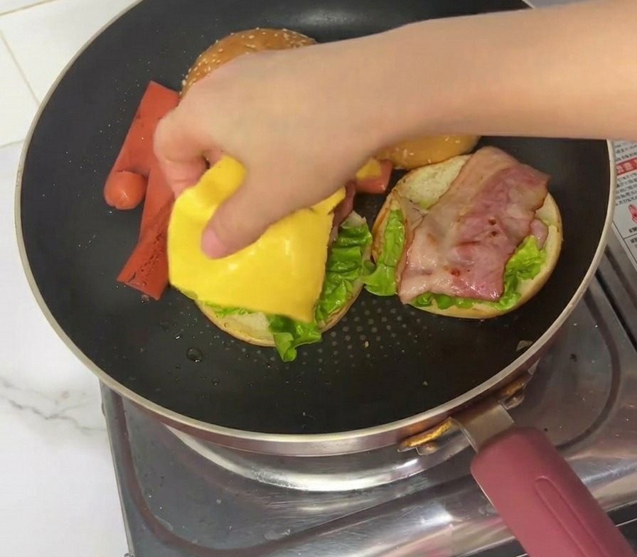 Une main plaçant une tranche carrée de fromage jaune sur lassemblage du burger dans la poêle.