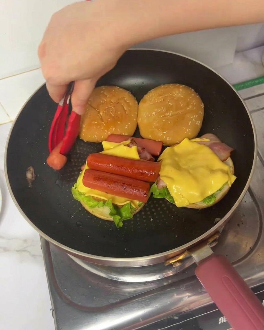 Une main utilisant des pinces pour placer un morceau dœuf plié sur le burger déjà garni.