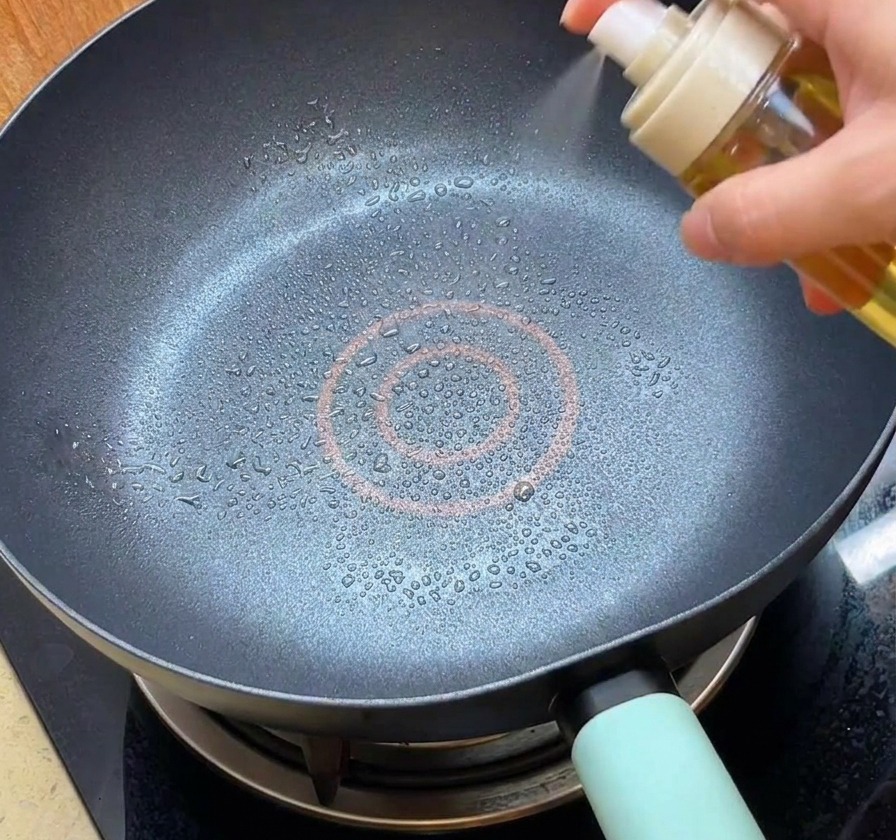 Une personne utilisant un vaporisateur blanc pour brumiser de lhuile dans une poêle antiadhésive noire propre.