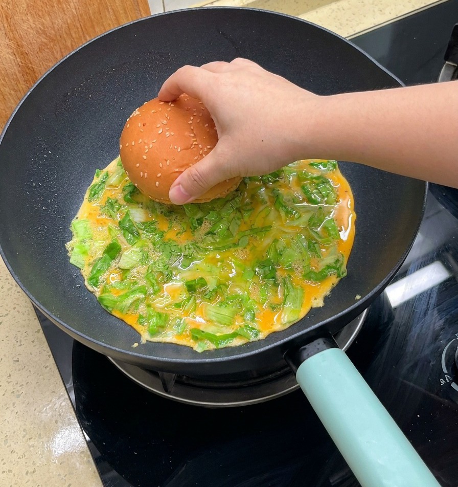 Une main plaçant un pain à burger au sésame sur une omelette à la laitue en cours de cuisson dans une poêle.
