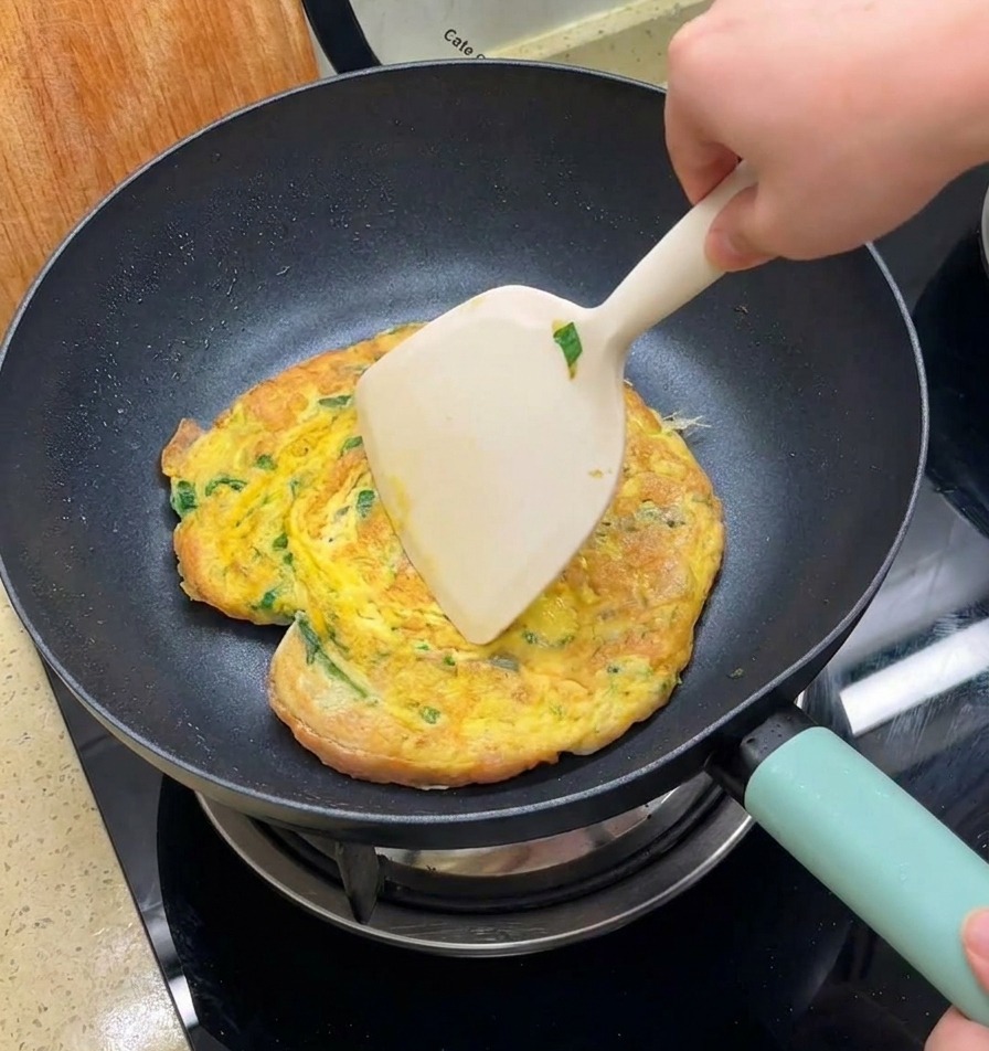 Une main utilisant une large spatule pour retourner une omelette dorée contenant de la laitue et des pains à burger dans une poêle noire.