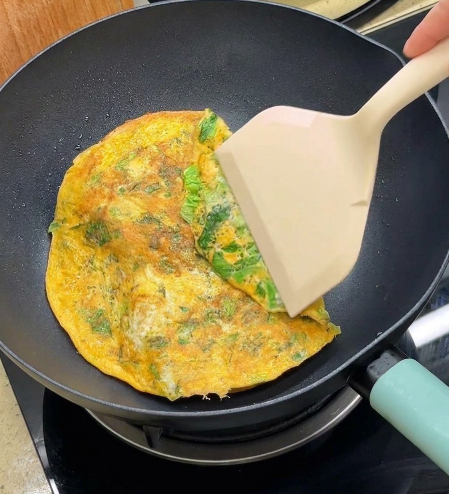 Une spatule repliant les bords dune omelette ronde sur deux pains à burger dans une poêle antiadhésive.