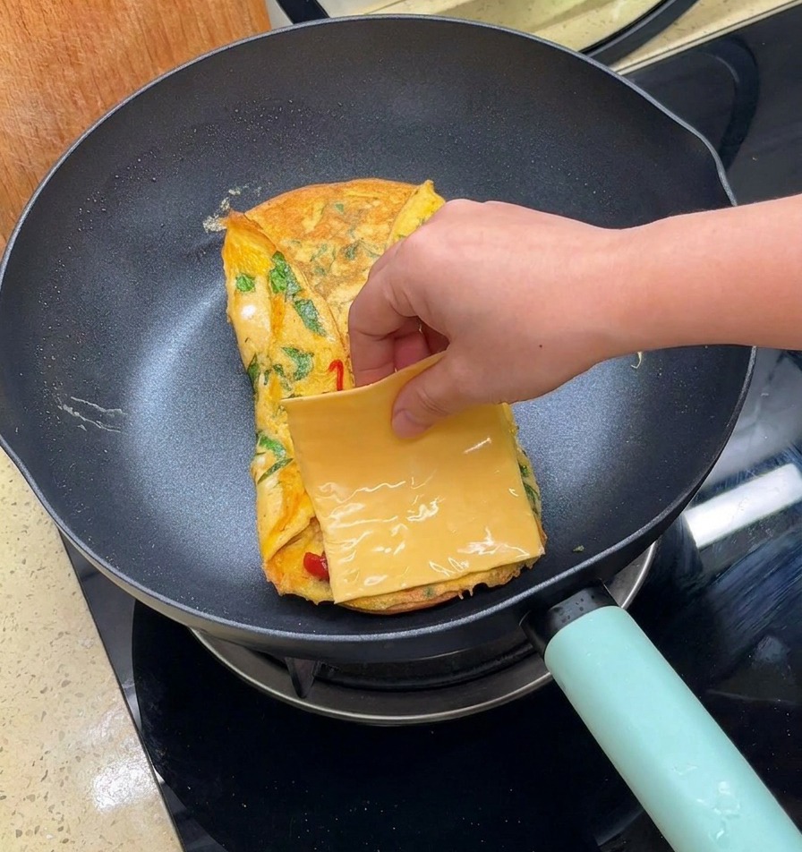 Une main plaçant une tranche carrée de fromage jaune sur une omelette pliée dans une poêle.