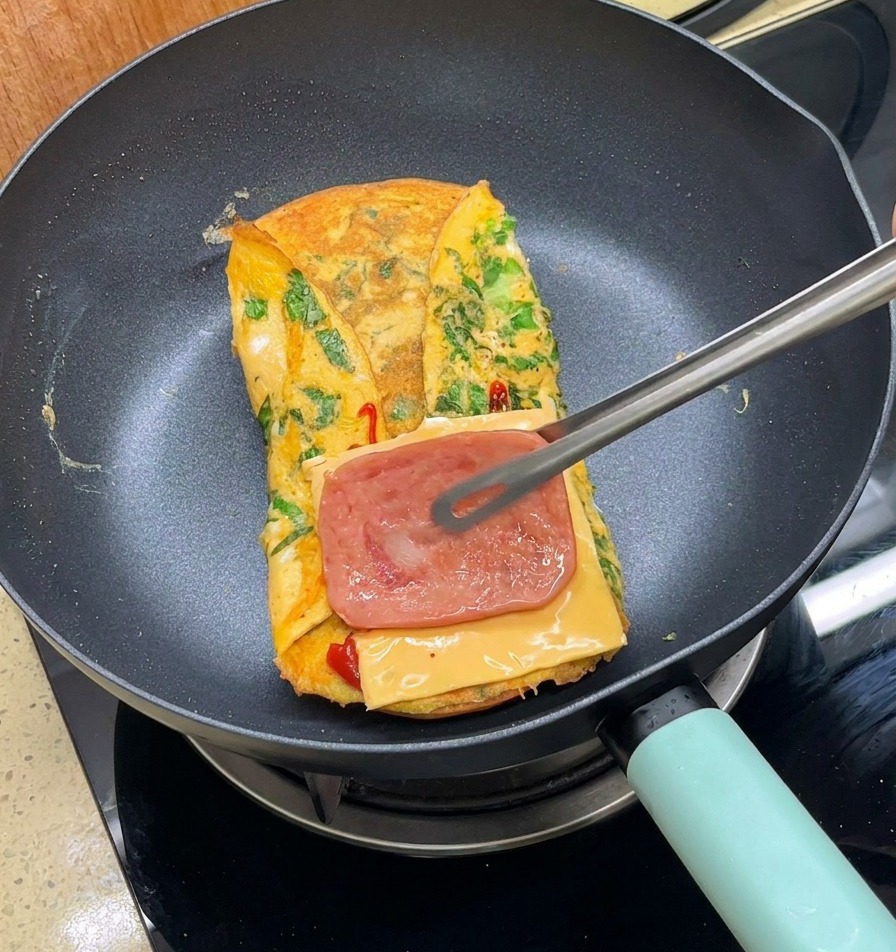 Des pinces métalliques plaçant une tranche de bacon cuit sur du fromage au-dessus dune omelette pliée dans une poêle.