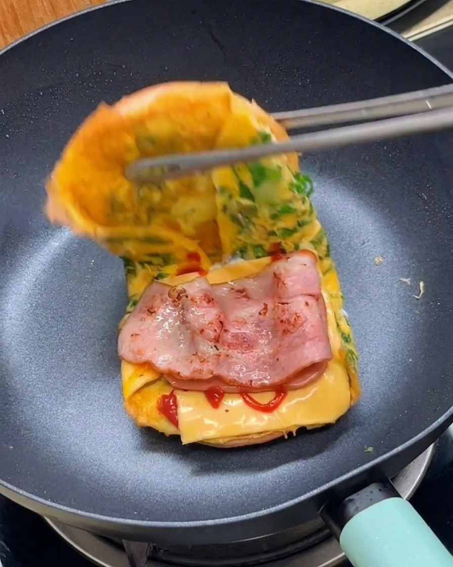 Des pinces pliant le bord dune omelette à la laitue pour finaliser un burger de petit-déjeuner dans une poêle.