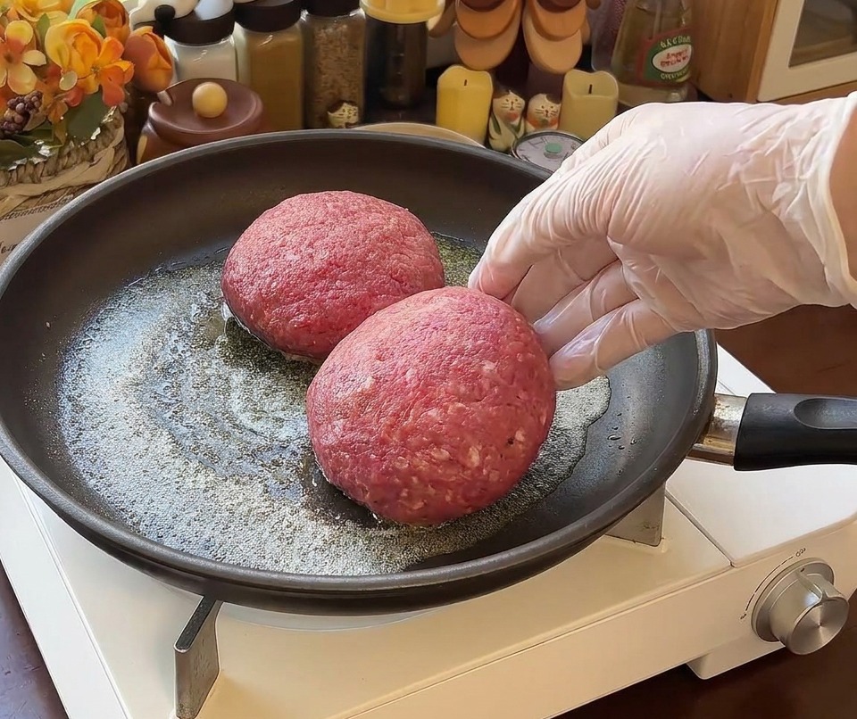 Deux épais steaks hachés de bœuf crus placés dans une poêle avec du beurre fondu, une main gantée visible sur la droite.