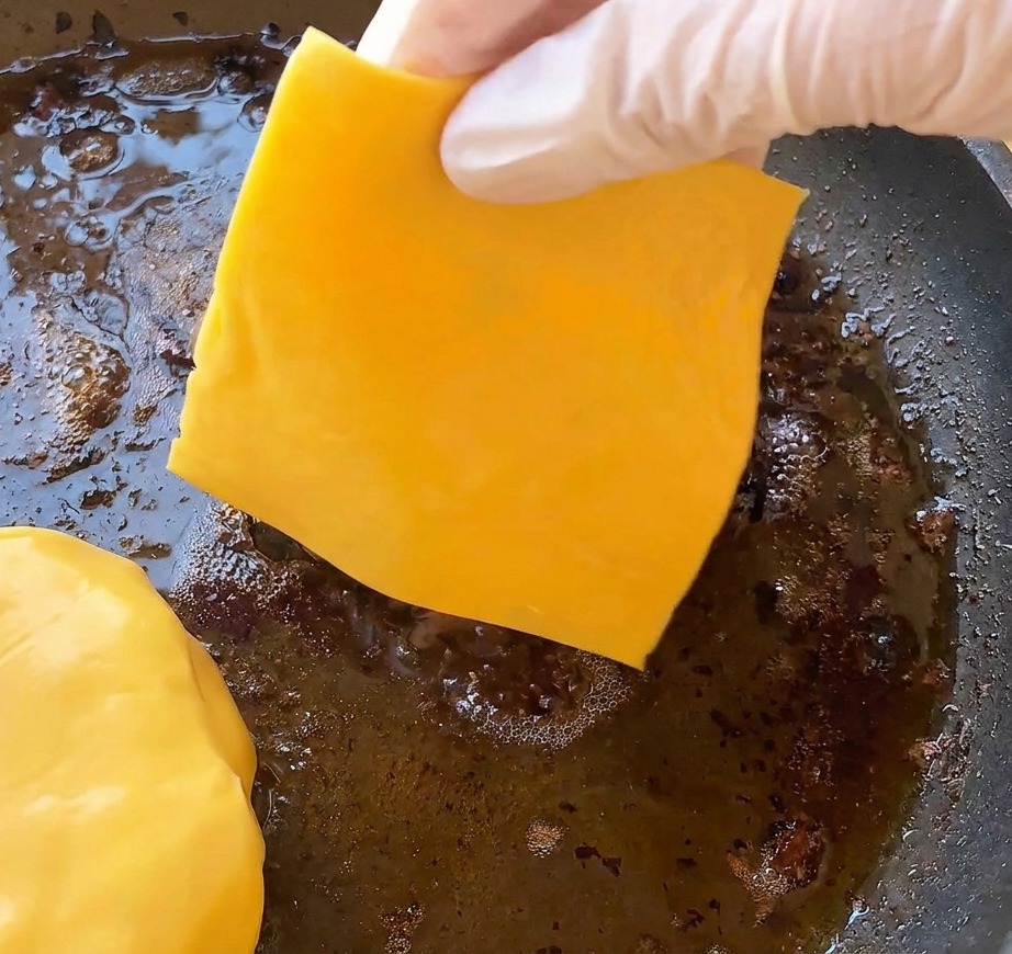 Une main gantée déposant une tranche de cheddar jaune vif sur un steak haché cuit grésillant dans une poêle.