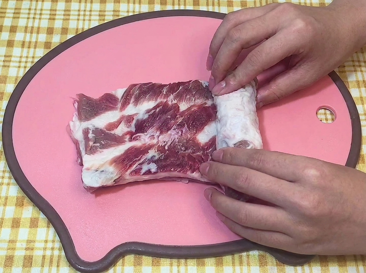Mains roulant fermement une pièce de poitrine de porc crue en cylindre sur une planche à découper rose.