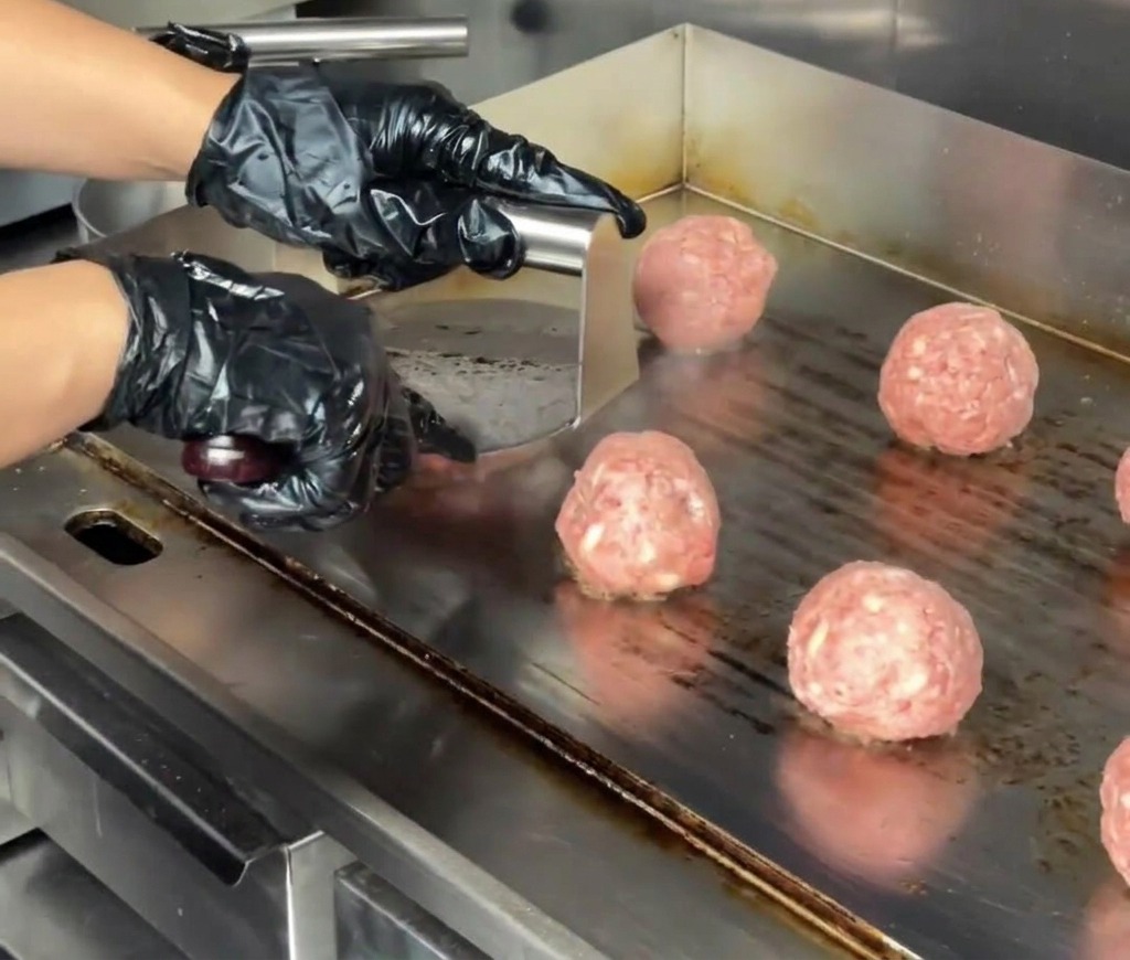 Gros plan dune presse à burger en inox aplatissant une boulette de bœuf en un steak épais sur une plancha professionnelle.