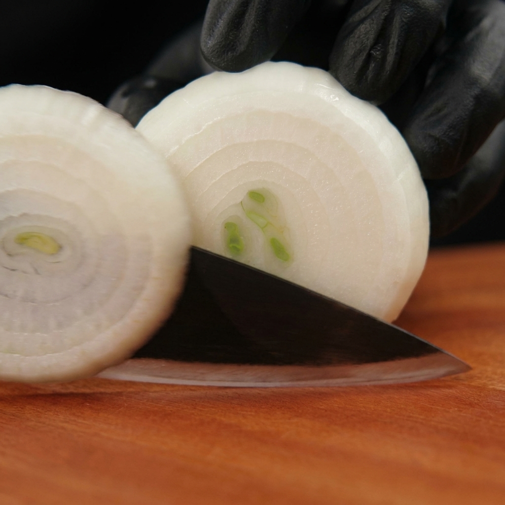 Mains gantées de noir utilisant un couteau pour couper un oignon blanc pelé en rondelles sur une planche en bois.