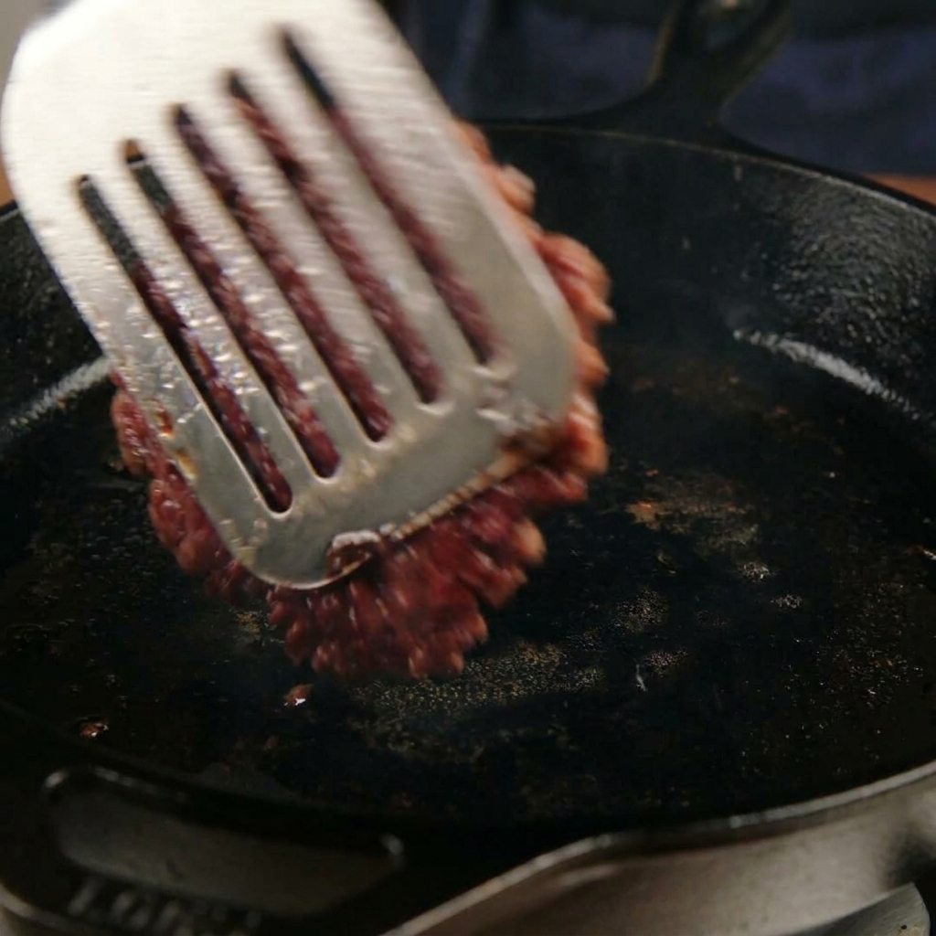 Retourner un steak de bœuf smashé et caramélisé dans une poêle avec une spatule en métal.