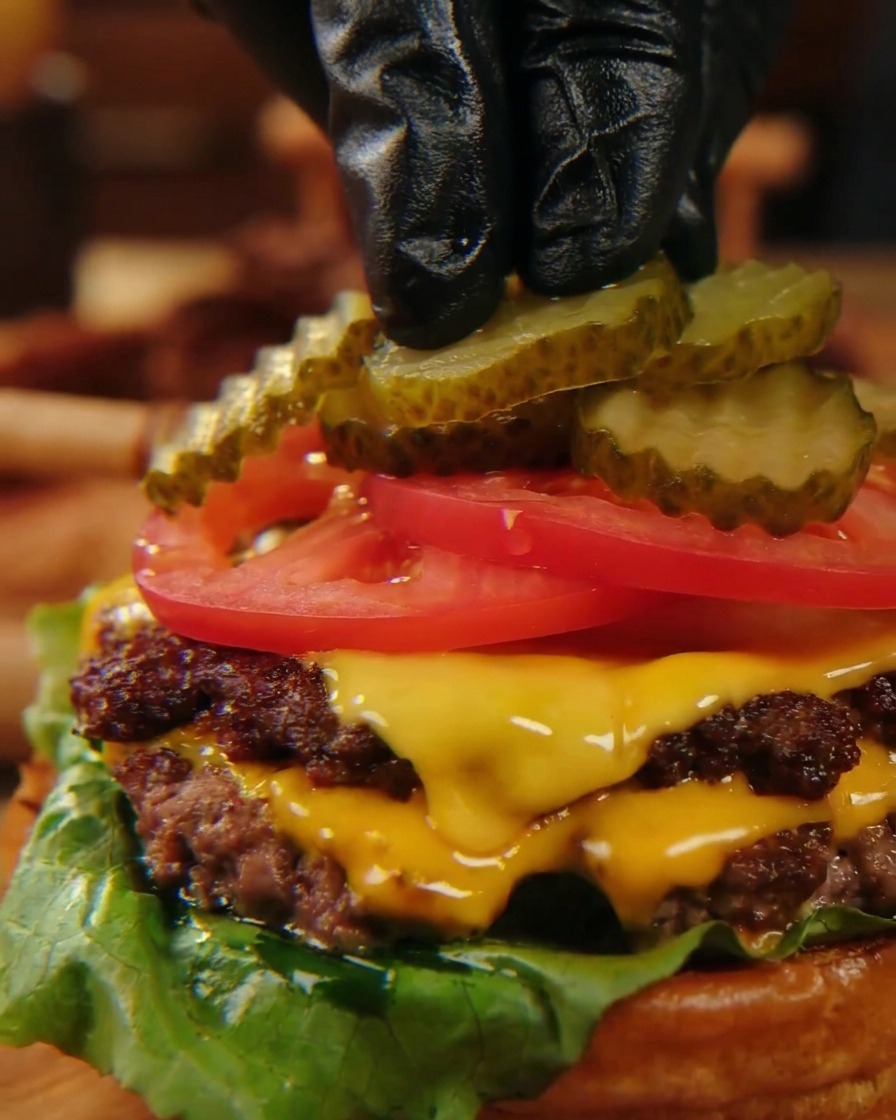 Des doigts gantés de noir plaçant délicatement des tranches de cornichons ondulés sur des tranches de tomates rouge vif au sommet dun cheeseburger empilé.