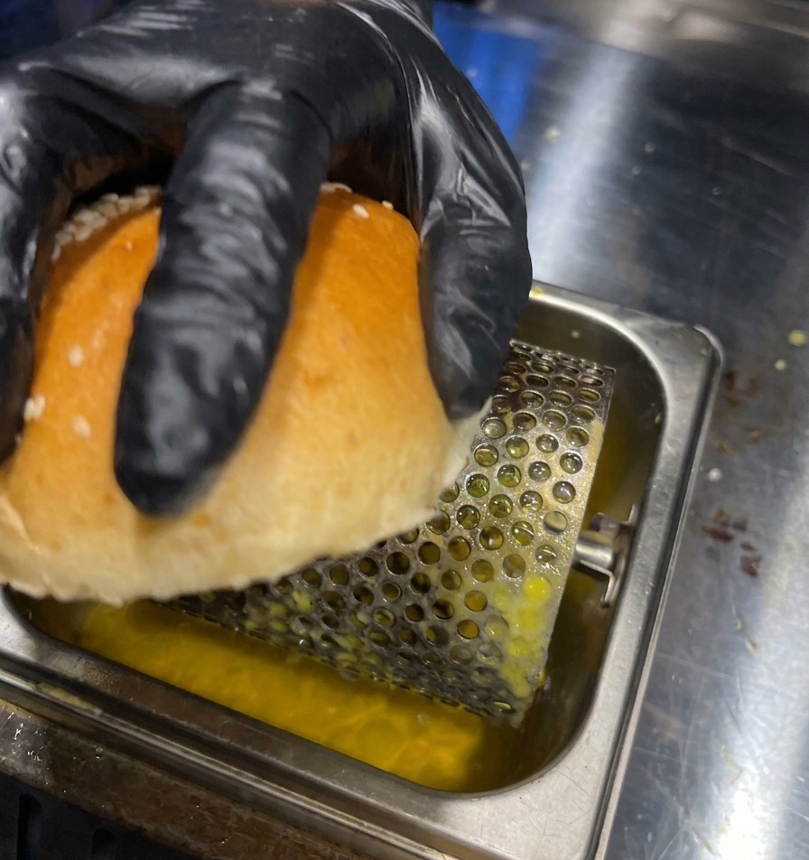 Une main gantée faisant rouler lintérieur dun pain burger sur un rouleau à beurre dans un bac en métal.