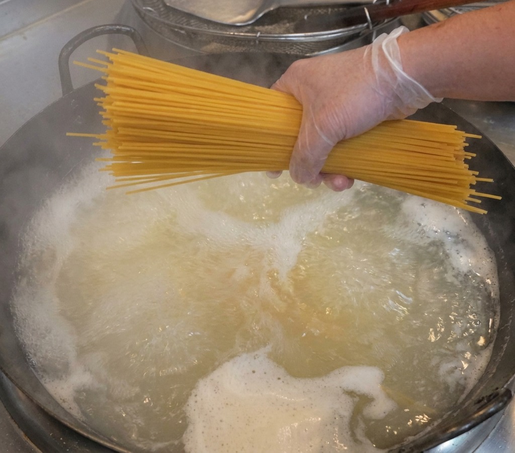 Une main gantée tenant un bouquet de spaghetti secs au-dessus dune casserole deau bouillante.