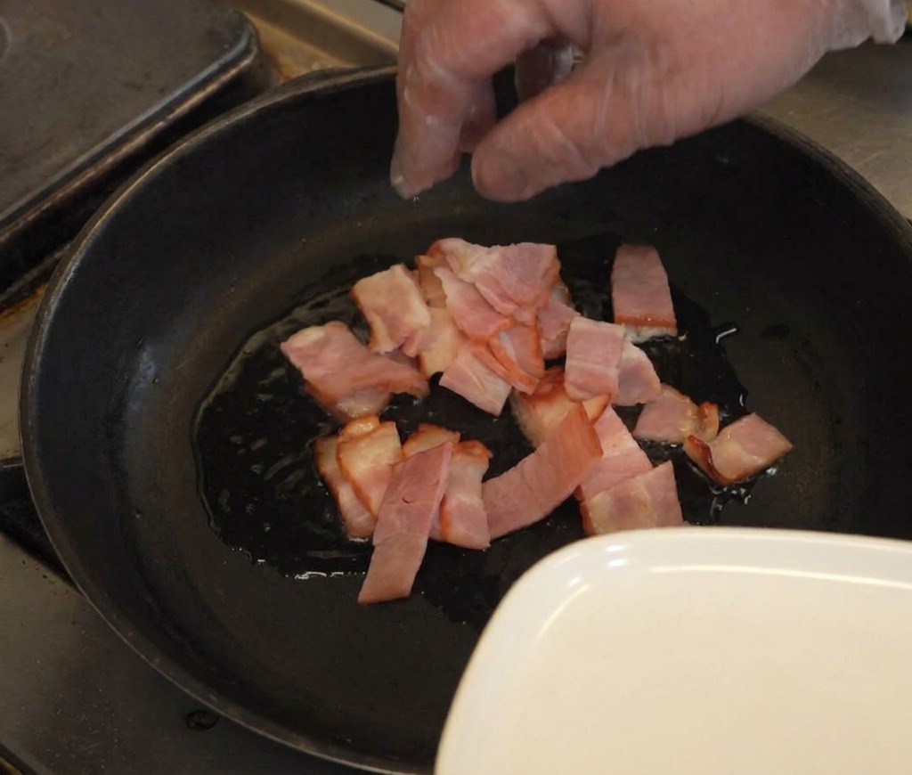Une main déposant des tranches de bacon cru dans une poêle sombre avec un léger filet dhuile.
