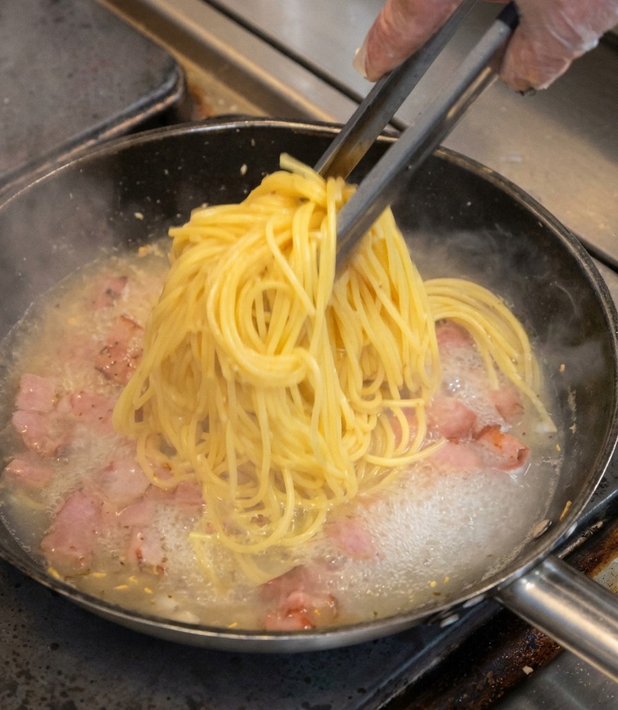 Un bouquet de spaghetti soulevé avec une pince et déposé dans la poêle avec le bacon.