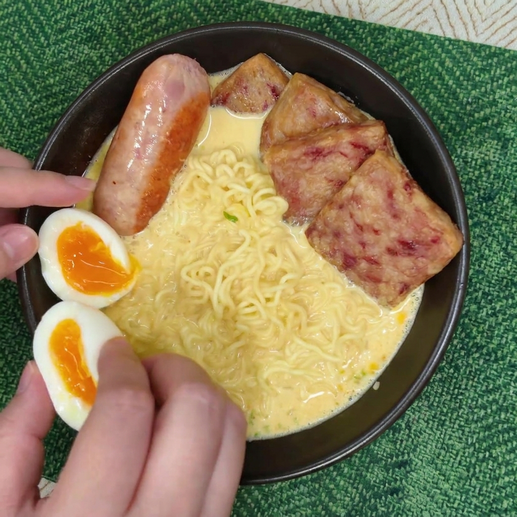 Des mains plaçant deux moitiés dœuf mollet au jaune orangé dans un bol de ramen à côté dune saucisse et de spam.