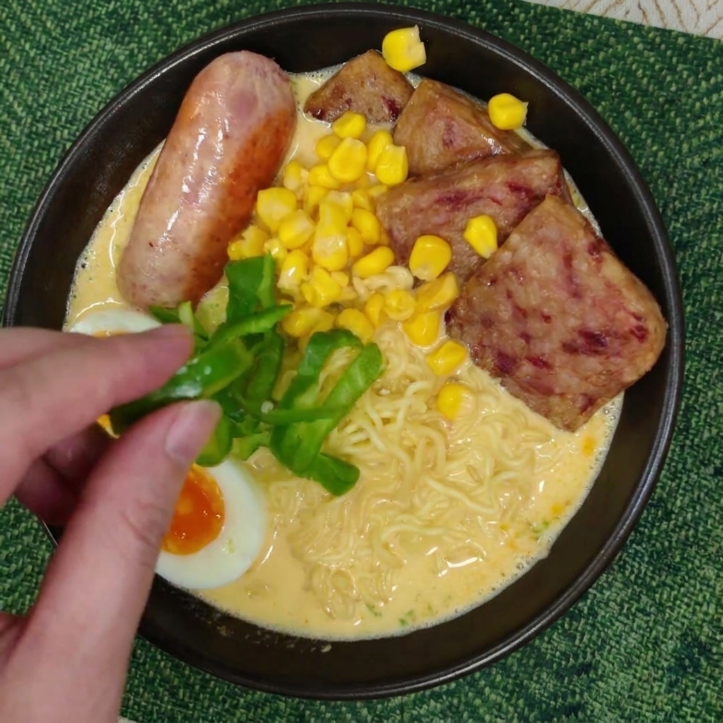 Une main plaçant des tranches de poivron vert frais sur le bol de ramen garni.