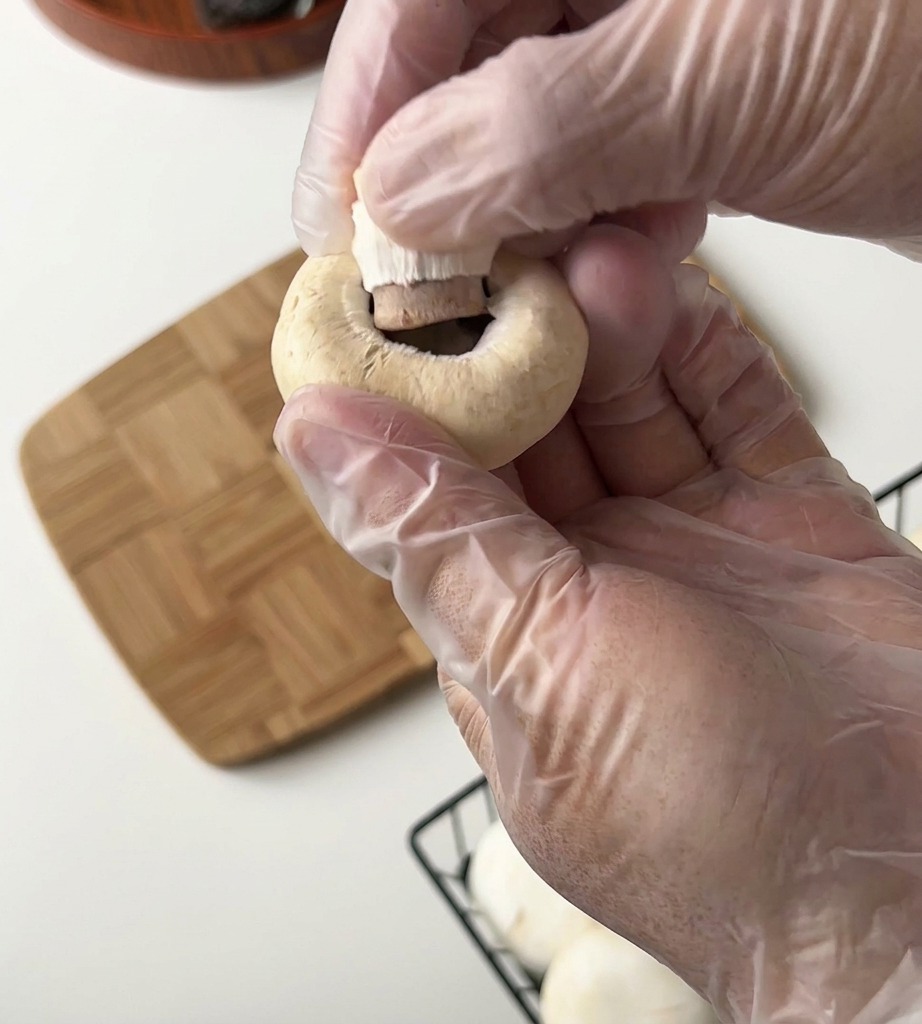 Une personne portant des gants en plastique retire la tige dun champignon de Paris au-dessus dune planche en bois.