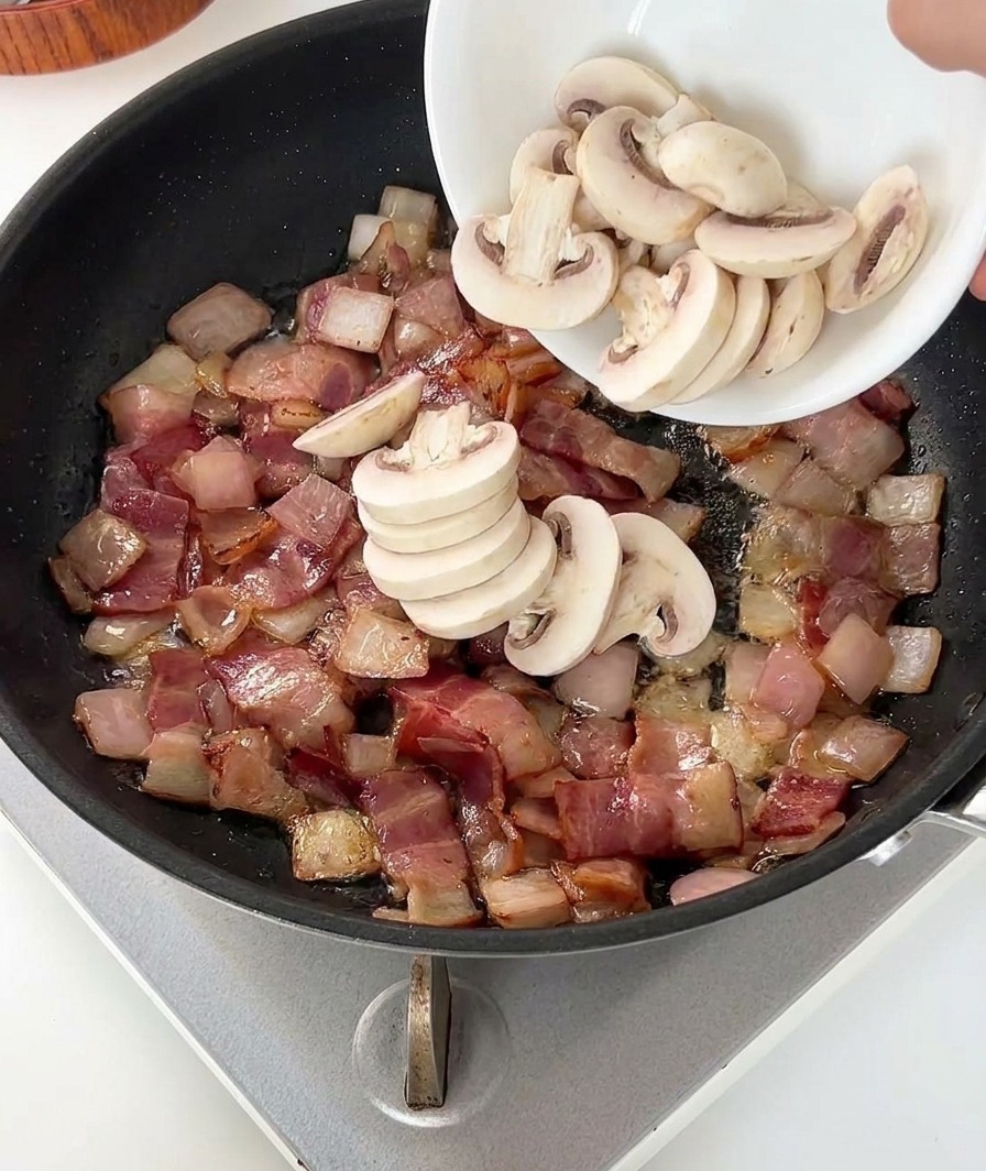 Une personne versant un bol de champignons blancs tranchés dans une poêle contenant du bacon doré et des oignons.