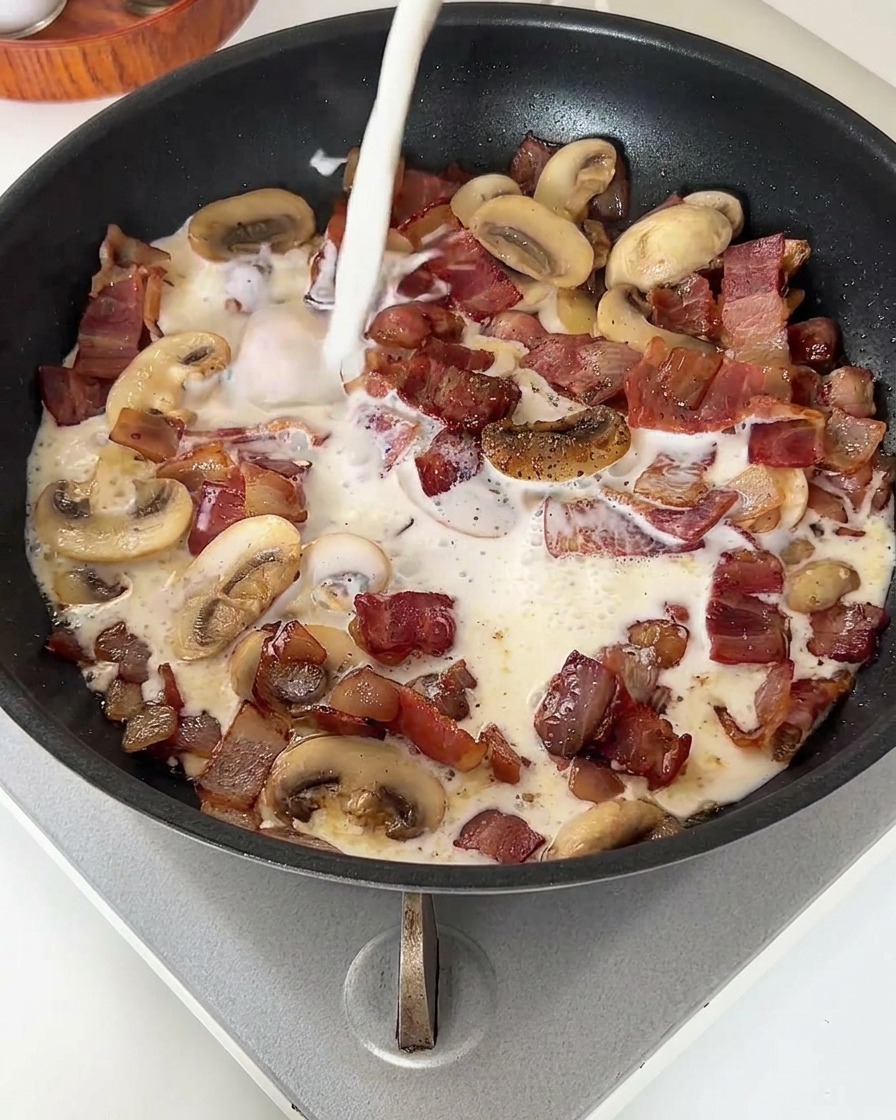 Un filet de lait blanc versé dans une poêle remplie de bacon et de champignons cuits.
