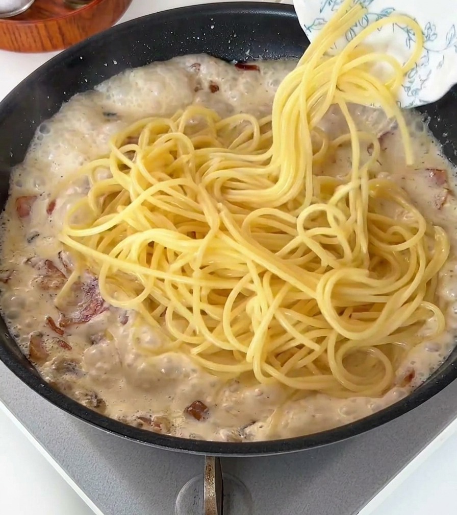 Spaghettis cuits déposés dans une poêle de sauce crémeuse bouillonnante avec du bacon.