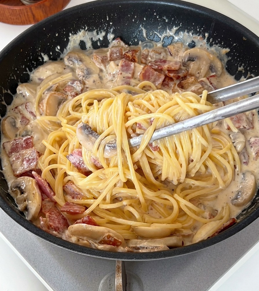 Une pince soulevant et mélangeant les spaghettis dans une sauce crémeuse épaisse avec du bacon et des champignons.