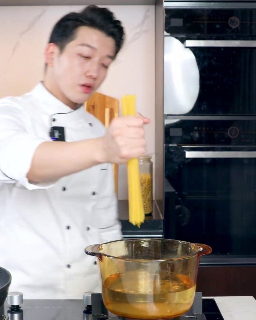 Un chef en uniforme blanc tenant un faisceau de spaghettis secs au-dessus dune casserole en verre remplie deau bouillante.