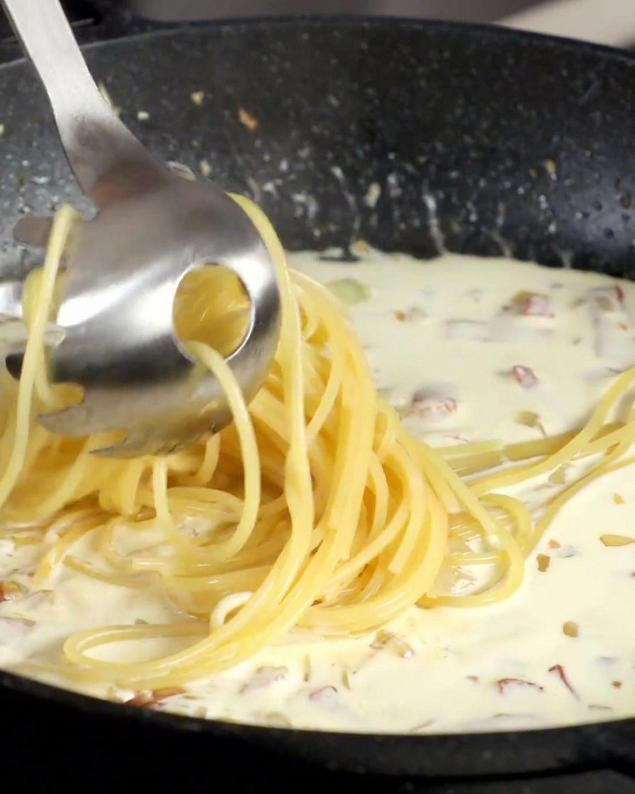 Une fourchette à pâtes soulevant des spaghettis cuits pour les déposer dans une sauce à la crème bouillonnante dans une poêle noire.