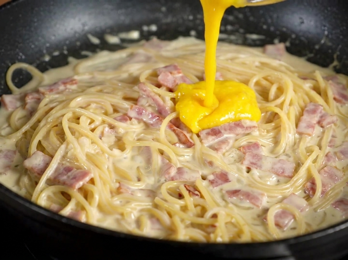 Un mélange épais de jaune dœuf et de fromage jaune vif versé sur des pâtes crémeuses et du bacon dans une poêle noire.