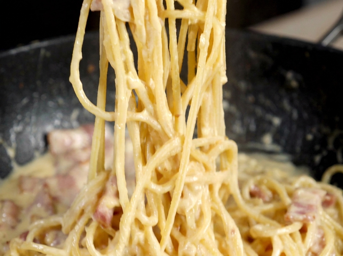 Un moulin à poivre en métal distribuant du poivre noir sur une poêle de pâtes façon carbonara.