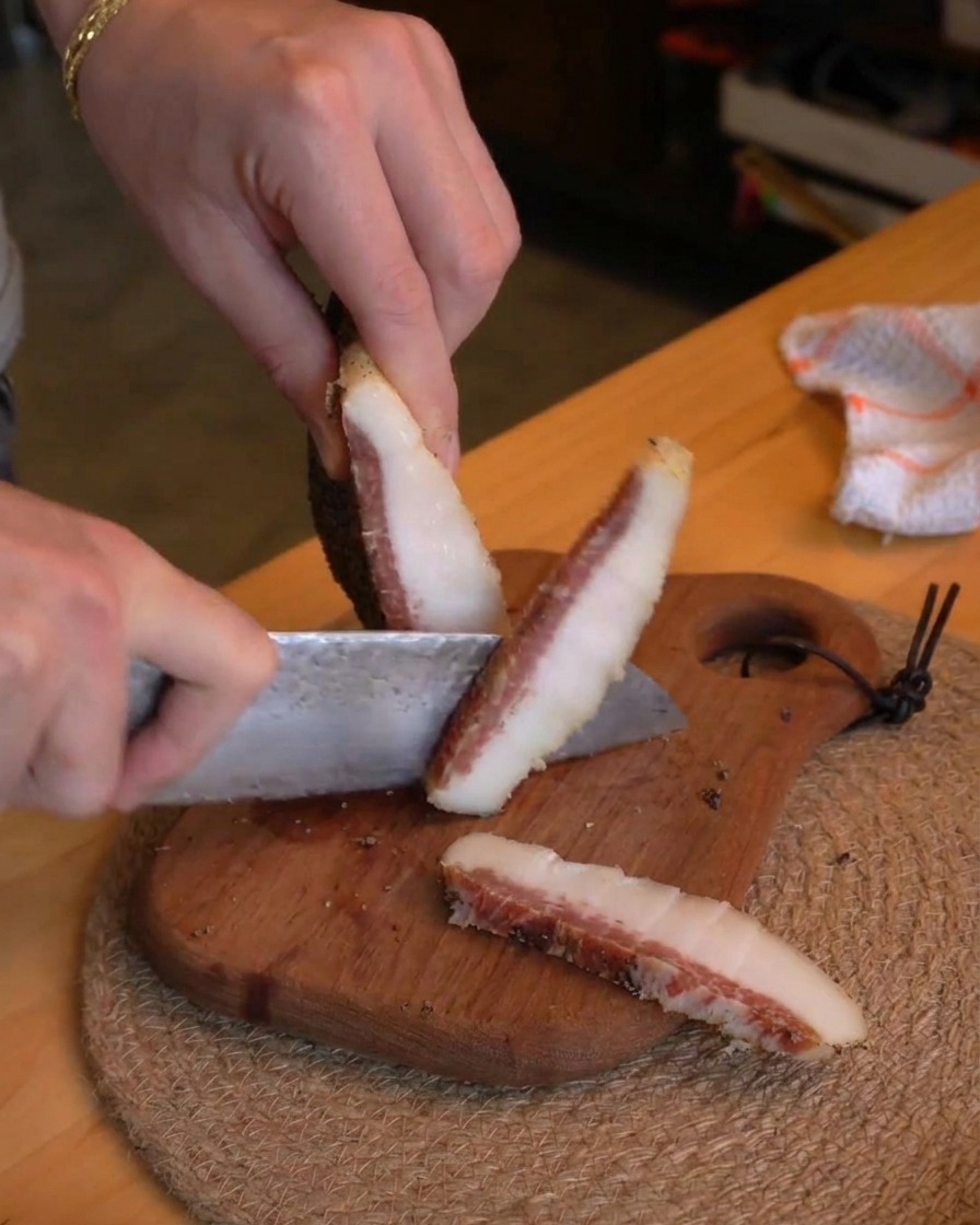 Une personne utilisant un couteau de chef pour couper dépaisses tranches de guanciale en petits bâtonnets sur une planche en bois.