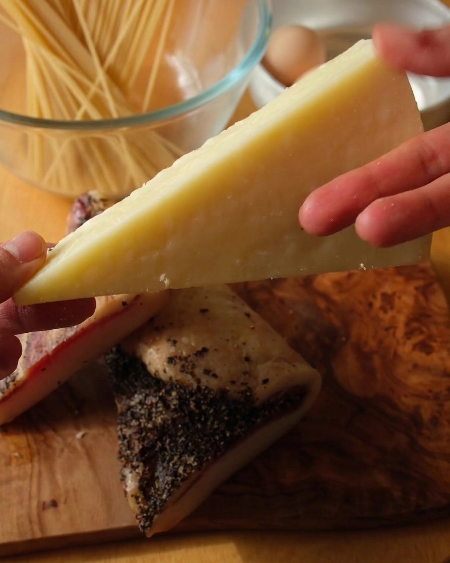 Gros plan dune main tenant un morceau de fromage Pecorino Romano pâle au-dessus de deux tranches épaisses de guanciale poivré sur une surface en bois.