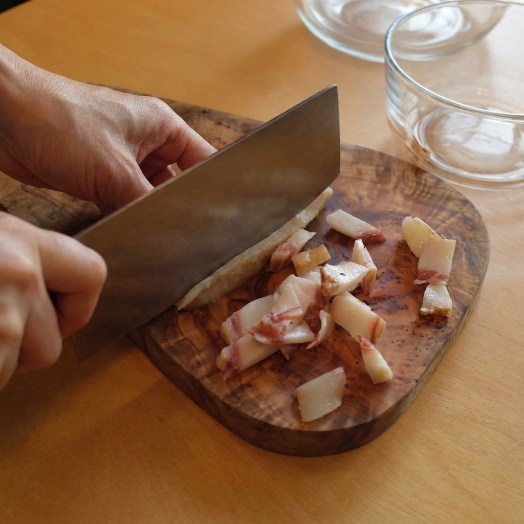 Une personne utilisant un grand couteau de cuisine pour couper des lanières de guanciale gras sur une planche à découper rustique en bois.