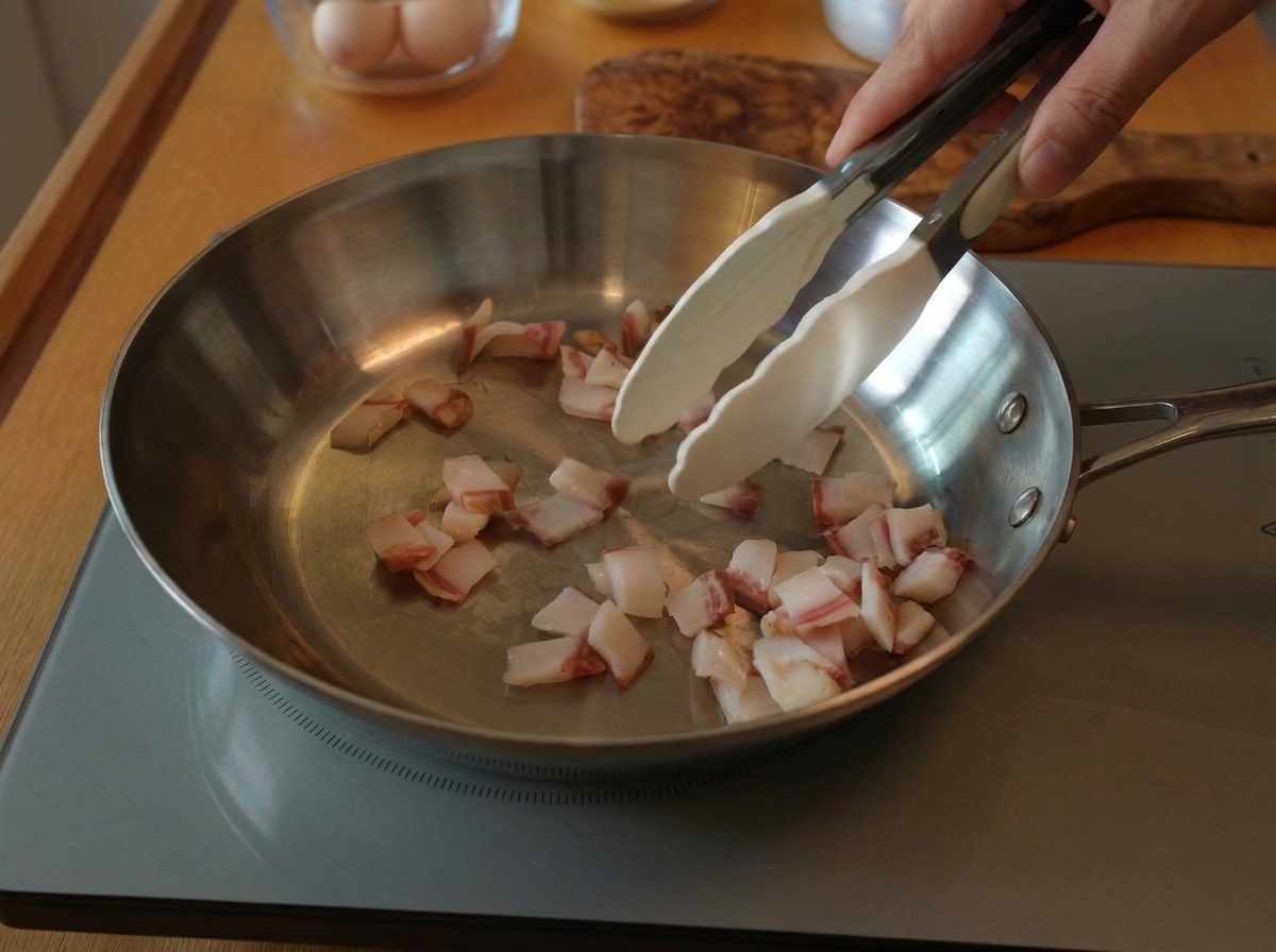 Une pince remuant des morceaux de guanciale cru dans une large poêle en inox sur une plaque à induction.