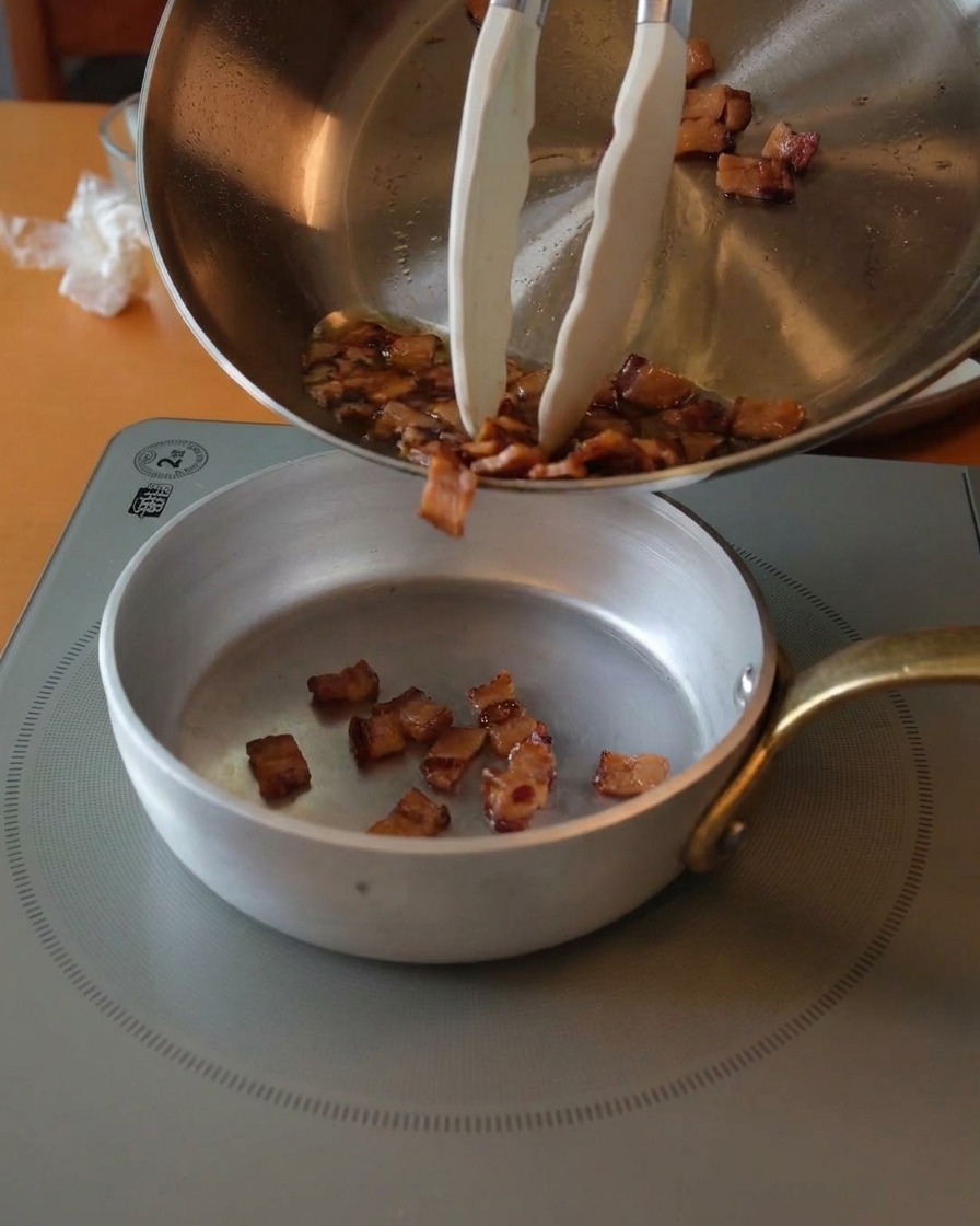 Morceaux de guanciale dorés et croustillants versés dune poêle en inox dans un petit bol argenté à laide dune pince.