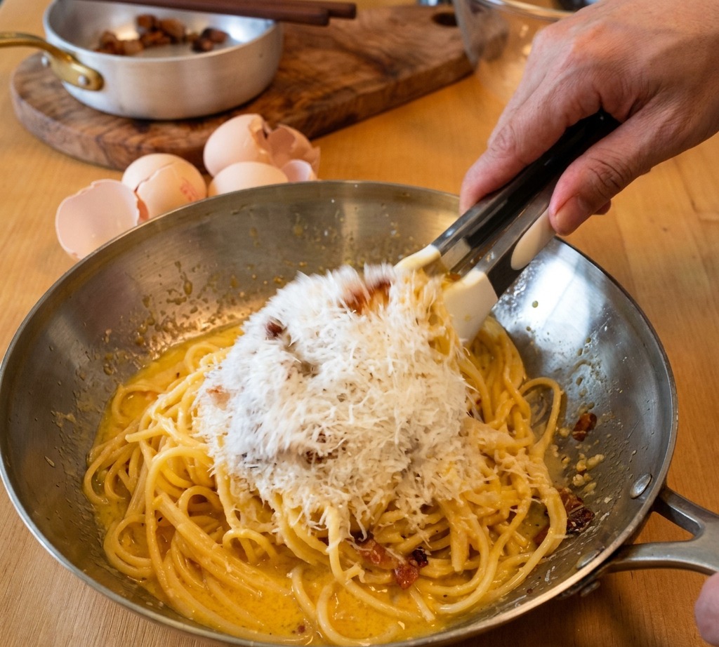 Pince mélangeant des spaghettis dans une poêle en métal pendant quun monticule de Pecorino Romano finement râpé est ajouté à la sauce crémeuse.
